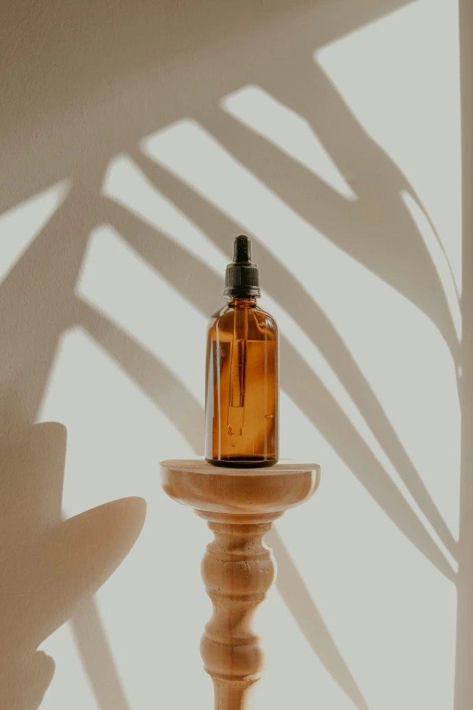 An amber glass dropper bottle on a wooden pedestal with palm leaf shadows